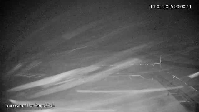 A dark and blurry image from a webcam shows an empty outdoor space, likely an airfield or runway at night. The foreground features a few indistinct shapes, possibly indicating structures or vehicles. Light streaks suggest movement, and there is minimal visibility overall. The timestamp indicates the image was captured on February 11, 2025, at 23:00:41. A logo for Leicestershire Aero Club is visible at the bottom left corner.