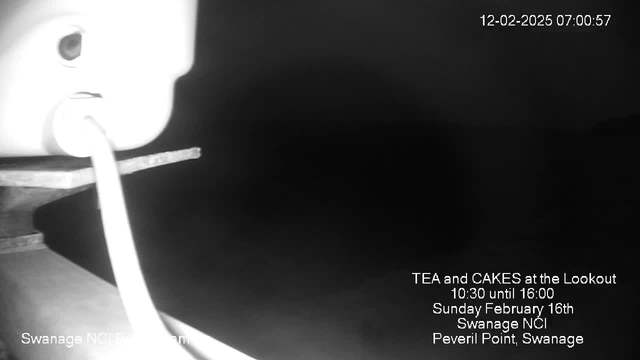 A dimly lit image showing the edge of a surface with a white cable extending towards the left. The background is dark and indistinct, suggesting it may be an outdoor or night scene. Text overlay at the bottom provides information about a tea and cakes event, including time and location details.