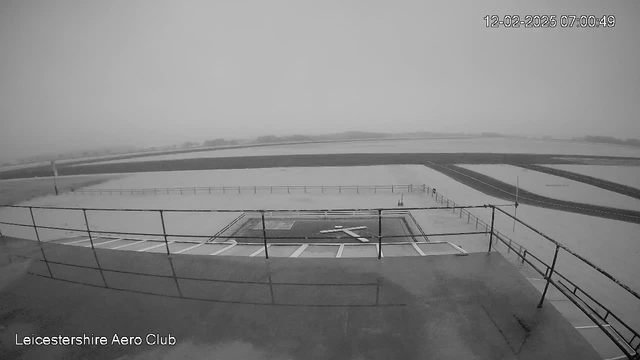 A black and white image from a webcam showing a view from a high vantage point. In the foreground, there is a landing strip marked with a white cross, surrounded by a low railing. Beyond the landing strip, the view extends to a large, flat area, possibly fields or an airport runway, with a hazy sky in the background. The date and time are displayed in the top right corner. The overall scene appears desolate and foggy, with limited visibility.