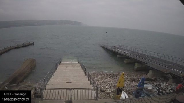 A coastal scene featuring a calm, gray sea under an overcast sky. In the foreground, there is a wide stone staircase leading down to the water, surrounded by railings. To the right, a wooden pier extends into the water, with a section of rocks visible below. There are two kayaks, one yellow and one blue, positioned on a rocky area at the edge of the water. The background includes a distant, blurred view of cliffs along the coastline, enhancing the tranquil atmosphere.