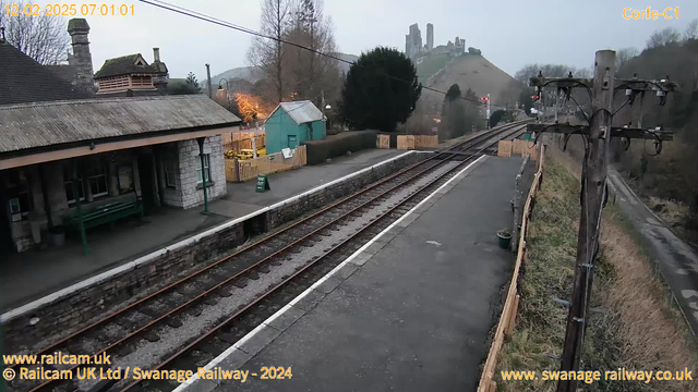 A view of a railway station with a stone platform and tracks. In the foreground, there is a green bench against a stone wall, and a sign indicating "WAY OUT." A small blue building is to the left, with a fence and some trees nearby. In the background, hills rise with ancient stone ruins visible on top, under a cloudy sky. The image is taken in early morning light.