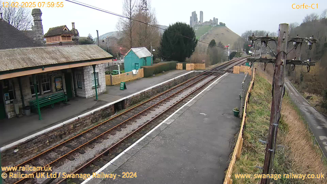 A view of Corfe Castle railway station in the morning. The scene shows a railway platform with a stone building covered by a sloped roof, featuring a green bench and a poster on the wall. To the left, there's a wooden fence with a sign indicating "WAY OUT." The railway tracks run parallel to the platform, leading off into the distance. In the background, a hill is visible with the ruins of Corfe Castle atop it. There are trees and a small green building behind the platform. A wooden utility pole with electrical wires stands on the right side of the image. The sky is overcast, and the overall atmosphere is calm and quiet.