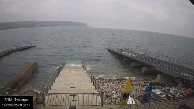 A view of a seaside area featuring a calm body of water extending into the horizon. On the left, there is a stone pier leading into the water. Adjacent to it are steps made of concrete leading down to the shore, which is rocky and has waves gently lapping at it. On the right, there is a wooden jetty extending into the water. Several colorful kayaks are parked along the edge, with a yellow one prominently displayed. The scene is under a cloudy sky, creating a muted light across the landscape. In the background, a distant cliff or coastline is visible.