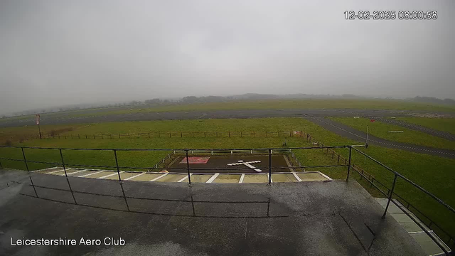 A foggy scene showing the Leicestershire Aero Club. The foreground features a railing and a portion of a flat roof. Below, there is an open grassy area with a small helipad marked by a large white cross. In the background, an unpaved runway can be seen extending across the landscape, with sparse trees and a low fence lining the perimeter. The sky is overcast and gray, contributing to a muted visibility throughout the image. The time stamp in the top right corner indicates the image was taken at 8:00 AM on February 12, 2025.