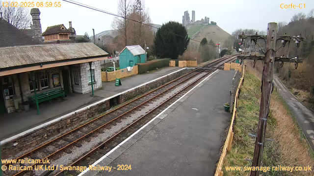 A railway station scene at Corfe, with a view of two railway tracks running parallel to the platform. The platform is made of stone with a wooden fence nearby. To the left, there is a stone building with a thatched roof, featuring a bench and a poster. In the background, a green wooden shed is visible, along with a sign indicating "WAY OUT." Beyond the station, a hill rises prominently, topped with the ruins of a castle, partially obscured by trees. The sky is overcast, and the scene conveys a quiet, rural atmosphere.