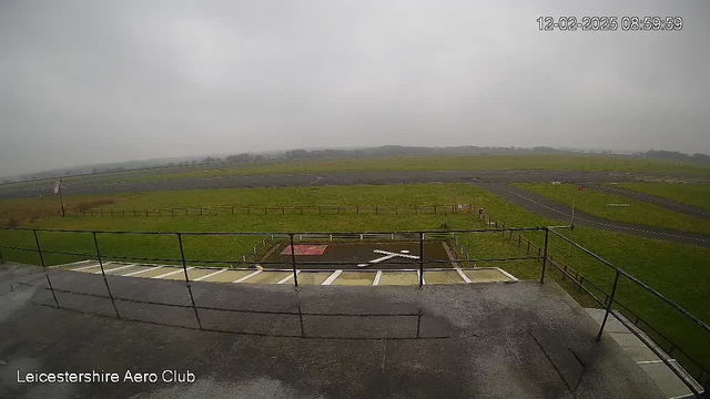 A view from a high vantage point overlooking a grassy airfield. In the foreground, a railing can be seen, along with markings on the ground resembling an "X". The airfield stretches out below, with several runways visible in the distance. The sky is overcast and gray, contributing to a dull and misty atmosphere. Fencing lines the perimeter of the airfield, and trees are faintly visible beyond the runway. The scene conveys a sense of quiet and stillness in a rural aviation setting.