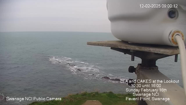 A view from a webcam overlooking a gray, cloudy sea. The water is calm with gentle waves approaching the rocky shore visible in the foreground. There are patches of rocks and a green grassy area leading up to the camera's base. In the background, a horizon meets the overcast sky, and at the bottom of the image, text promotes an event called "Tea and Cakes at the Lookout" with details about the date and location.