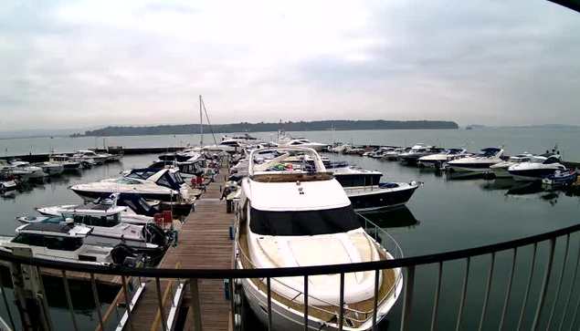 A wide view of a marina featuring multiple boats lined up at a dock. The scene is set under a cloudy sky, with water reflecting the overcast light. Various vessels are parked, including a large white yacht in the foreground and smaller boats in the background. A wooden walkway runs alongside the dock, and a distant shore is visible across the water.