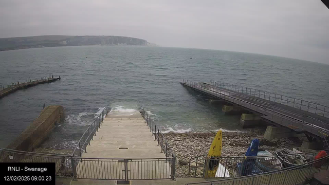 A coastal scene showing a stone stairway leading down to the water, flanked by a metal railing. To the right, two kayaks in bright yellow and blue are stored on a platform. In the background, a rugged coastline can be seen, with gray skies above. The water is choppy with gentle waves, and a wooden jetty extends into the sea on the left side of the image.