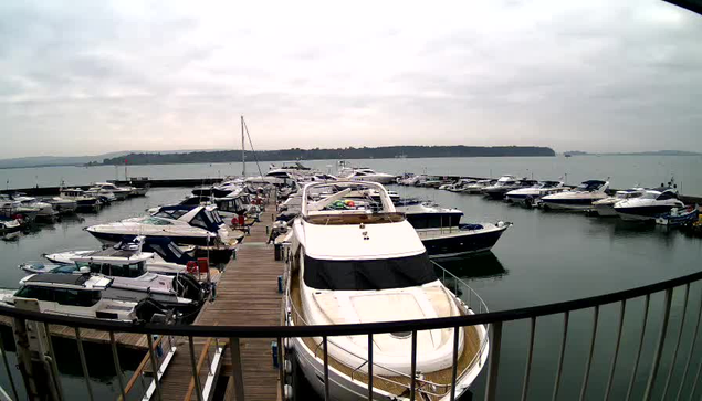 A view of a marina on a cloudy day, with numerous boats docked in the water. Several yachts and smaller vessels are visible, arranged neatly along wooden docks. The water reflects the overcast sky, and a distant shoreline is faintly seen in the background. The scene conveys a calm, maritime atmosphere.