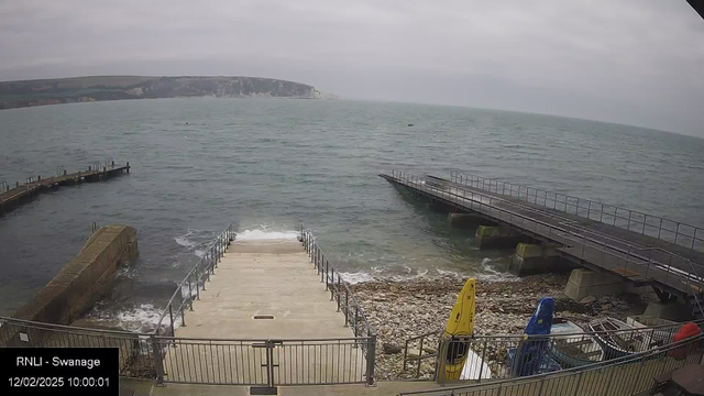 A view of the coastline at Swanage on a cloudy day. In the foreground, there is a set of stone steps leading down to the water, with gentle waves lapping at the base. To the left, a small jetty extends into the sea. Three colorful kayaks—a yellow one, a blue one, and a red one—are visible on the right side. The horizon features cliffs in the distance, shrouded in mist, under a gray sky.