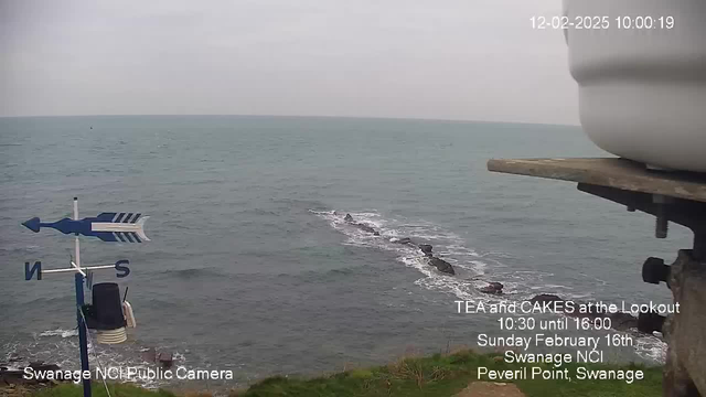 A coastal scene featuring the sea and a rocky shoreline. In the foreground, there is a weather vane pointing North, with a blue arrow and other directional indicators. The sky is overcast with gray clouds, and the ocean appears calm with gentle waves lapping at the rocks. The date and time are displayed in the top right corner, along with text advertising a tea and cakes event at a lookout point in Swanage.