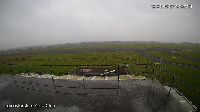 A view from a webcam overlooking a green field and an airstrip. The sky is overcast, and the scene appears cloudy and gray. In the foreground, there is a railing, and to the left, a small helipad with an "X" marking. Beyond the railing, a grassy area stretches out towards an airstrip lined with sparse fencing. The area is empty, with no visible aircraft or people. A slight road is visible in the distance, along with some trees and a few structures farther away.