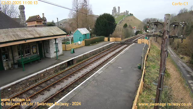 A railway station scene featuring a platform with a stone-built shelter and a green bench. In the background, there is a small green building and a wooden fence enclosing a garden area. The railway tracks extend into the distance. A prominent feature in the background is a hill with a castle ruin on top. The sky is overcast, and the surroundings include trees and a power pole with wires. The image has a timestamp indicating the date and time.