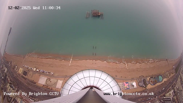 A high-angle view of a beach and ocean, captured from a webcam. The camera is pointed downwards, showing a sandy beach with a few wooden structures and a pier visible in the water. The coastline features a mix of sand and some small waves. To the right, there are amenities and a swimming pool near the beach. The sky is slightly overcast, and timestamps show the date and time in the top left corner. The logo "Powered By Brighton CCTV" is displayed at the bottom.