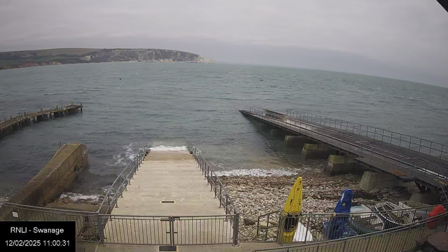 A rocky shore leading to water is visible in this webcam image. There are two piers extending into the sea, one wooden and one concrete. A set of stairs leads down to the water, bordered by a metal fence. On the right, there are three boats resting on the ground, with one yellow kayak prominently displayed. The sea is slightly choppy, and distant cliffs can be seen in the background under a cloudy sky. The date and time of the image are displayed in the bottom left corner.