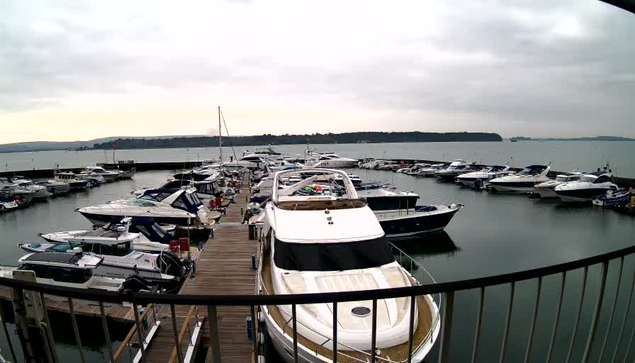 A marina filled with various boats and yachts docked in the water. The boats are arranged in a semicircle around a wooden dock. The sky is overcast with gray clouds, and the water appears calm. In the background, there are hills and trees along the shoreline. The scene conveys a tranquil waterfront setting.