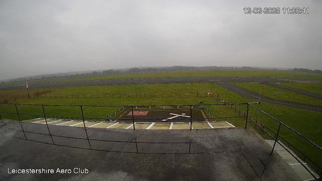 A view from a webcam at Leicestershire Aero Club, showing a flat, grassy airfield under a cloudy sky. In the foreground, there is a railing and a concrete surface. In the background, a large expanse of green grass is visible, with a paved runway crossing horizontally. A small area with a red marking and a white 'X' symbol is located near the bottom of the image, indicating a landing or takeoff point. The atmosphere appears overcast with low visibility.