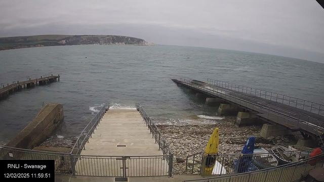 A view of a coastal area featuring a wooden pier extending into the sea on the left side, with a staircase leading down to a rocky shoreline in the middle. There are kayaks in yellow and blue positioned on the right side. The ocean appears calm with gentle waves and a cloudy sky in the background, revealing cliffs along the shore in the distance.