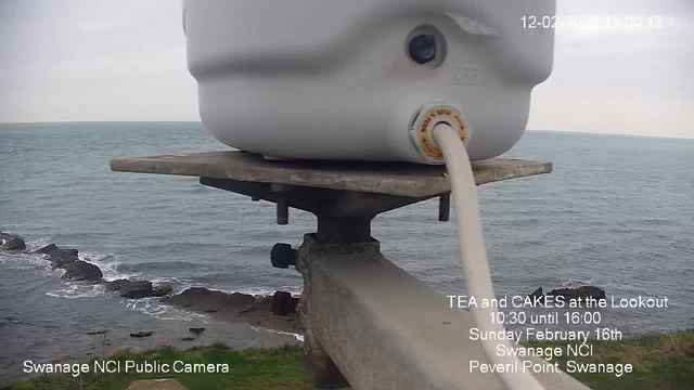 A white container is positioned on a metal platform overlooking the sea. In the background, the calm waters of the ocean meet a cloudy sky. Rocks are visible along the coastline, and patches of green grass are in the foreground. Text in the lower part of the image indicates a public camera in Swanage, with details about a tea and cakes event on February 16th.