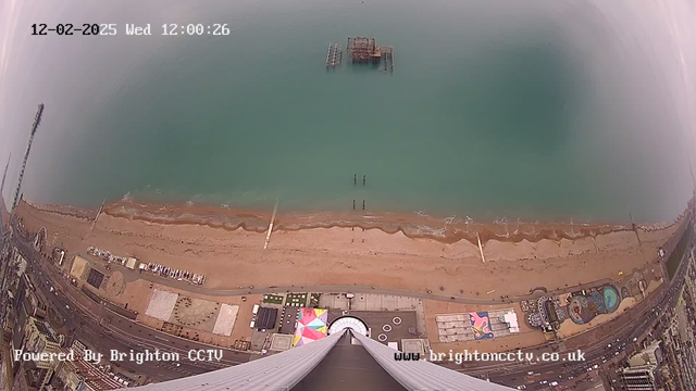 A high-angle view of a beach and ocean. The sandy shore is visible, lined with gentle waves. An old pier structure juts out into the water, and several poles extend from the water's edge. Below, colorful structures and roads are visible, with cars parked along the edge. The scene is somewhat cloudy, and the date and time are displayed in the upper left corner.