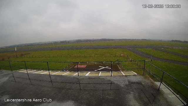 A view from a webcam located at Leicester Aero Club, showing an expansive grassy area under a cloudy sky. In the foreground, there's a railing and what appears to be a concrete platform with a white geometric design resembling an 'X'. Beyond the platform, a runway is visible, marked by parallel black lines, with a grassy field on either side. The surroundings are primarily green, with a few fences along the edges and the landscape fading into the distance. The image is taken during overcast weather, with no signs of activity.