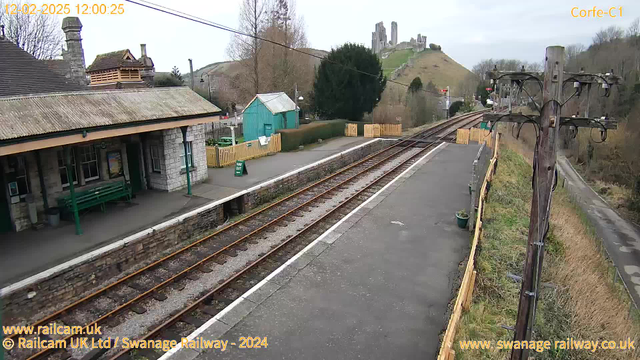 A railway station scene with a stone building featuring a peaked roof and a canopy. There are wooden benches under the canopy. The station has a green sign that reads "WAY OUT." A row of tracks runs through the foreground, leading to a distant view of a hill with ruins on top. To the left, there is a small green shed and a fence. The background shows trees and a cloudy sky.