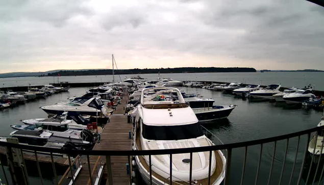 A marina filled with multiple boats and yachts docked at a wooden pier. The water is calm and reflects the cloudy sky overhead. There are several boats of various sizes, and a distant shoreline is visible in the background. The scene appears peaceful, with no people present.