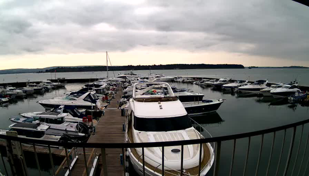 A marina scene with multiple boats docked in calm water. Prominent in the foreground is a large white yacht with a raised bridge. Surrounding it are smaller boats of various sizes and colors, predominantly white and blue, moored along a wooden pier. The sky is overcast with gray clouds, and distant hills are visible along the horizon. The overall atmosphere is tranquil and serene.