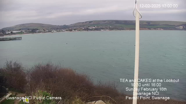 A coastal view from a lookout point. The image shows a calm sea with a light turquoise color, dotted with a few buoys. In the background, there are buildings along the shore and rolling green hills. A wooden pier extends into the water, partially visible on the left side of the image. On the right side, a white flagpole stands with no flag. The sky is overcast, adding a gray tone to the scene. Text overlay provides details about an event featuring tea and cakes at the lookout, scheduled for February 16th.