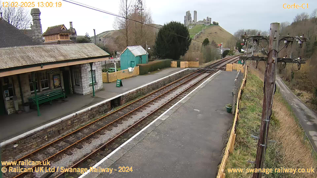 A railway station platform viewed from an elevated angle. On the left, there is a stone and wood building with a sloped roof and large windows. A green bench is visible on the platform. The platform is bordered by a low stone wall and there are railway tracks running along the foreground. In the background, a hillside with ruins of a castle is visible, surrounded by trees. A green structure, possibly a shed, is situated on the left side beyond the station.