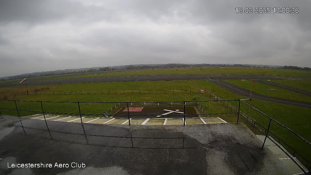 A view from a webcam at the Leicestershire Aero Club, showing a large, grassy field under a cloudy sky. In the foreground, there is a railing terrace with a few steps leading down. The runway is visible, along with a small area marked with a red sign. The landscape is mostly flat, with a few distant trees and structures in the background. The atmosphere is overcast, with muted lighting.