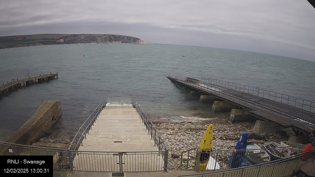 A coastal scene featuring a partially cloudy sky. In the foreground, there is a concrete ramp leading down to the water, bordered by a metal railing. To the left, a wooden pier extends into the water, while to the right, a larger modern pier stretches out further. The water appears calm with a slight ripple, and there are several colorful kayaks parked on the shore, including yellow and blue ones. In the background, cliffs and greenery create a scenic landscape along the coast.