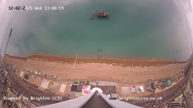 A view from a high vantage point overlooking a beach and ocean. The sandy beach stretches across the lower part of the image, bordered by calm turquoise water. A few structures, including a pier with ruins, are visible in the water. On the beach, there are scattered objects and signs of activity, with colorful structures and patterns seen on the land. The sky appears overcast, hinting at a cloudy day. In the corner, a timestamp shows the date and time of the image.