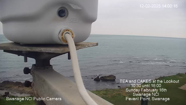 A grayish view of the sea under a cloudy sky is captured from a high perspective. In the foreground, a white container is mounted on a metal structure, with a cable extending from it. Below, a rocky shore is visible, with scattered boulders along the waterline. The scene is part of a public webcam feed, and text in the bottom left corner identifies it as "Swanage NCI Public Camera." Additional text promotes an event featuring tea and cakes at a lookout on February 16th.