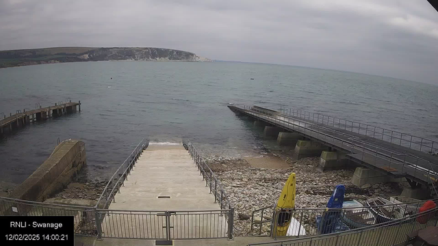 A view of a coastal scene featuring calm water under a cloudy sky. On the left, there’s a wooden pier extending into the water, and to the right, a concrete ramp leads down to the water’s edge, with rocky shorelines visible. In the foreground, yellow and blue kayaks are positioned next to the ramp, along with some stacked boats. The background showcases a rocky cliff and green hills.