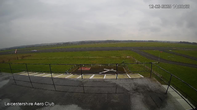 A view from a webcam at Leicestershire Aero Club shows an expansive airfield under a cloudy sky. In the foreground, there is a gray rooftop with a railing, and a part of the ground marked with yellow and white strips. In the distance, a grassy area is visible alongside a runway where a small aircraft is parked. Fences delineate the area, and a windsock can be seen on the left side, indicating wind direction. The overall scene has an overcast ambiance. The time stamp in the corner indicates the image was taken on February 12, 2025, at 14:00:22.