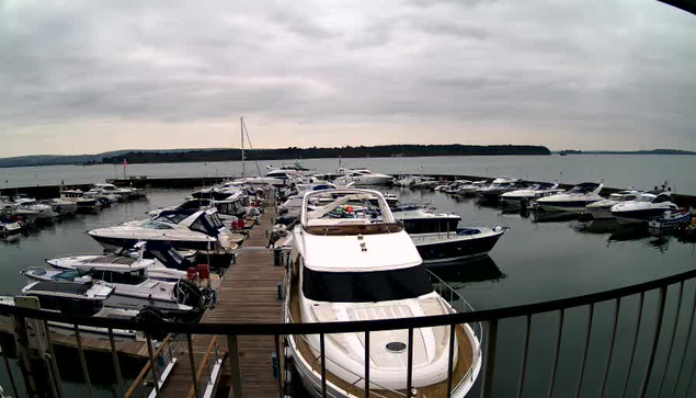 A marina scene on a cloudy day, with numerous boats moored at a dock. The water is calm, reflecting the gray sky. Several yachts and smaller boats are visible, with some docked side by side. A wooden walkway leads from the viewer's perspective into the marina. In the background, rolling hills can be seen along the shoreline.