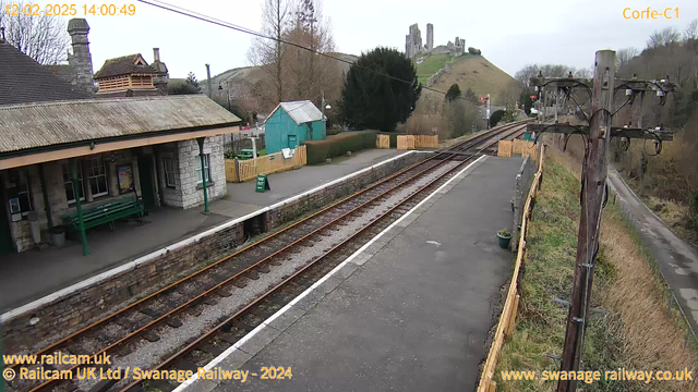 A railway station is depicted, featuring a stone platform with a green bench and a ticket booth area. To the left, there is a wooden structure with a pointed roof. The background showcases rolling hills and a castle ruin perched on the hillside. Several railway tracks run through the foreground, leading off into the distance. A wooden fence lines the platform on the right, which also has a sign indicating a way out. The sky is overcast, creating a grey atmosphere.