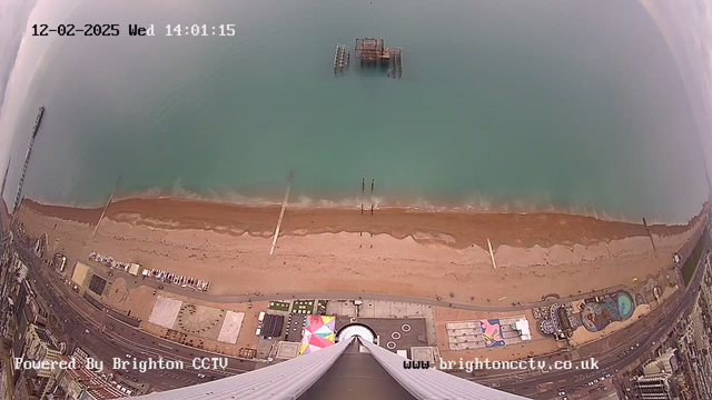 A wide aerial view of Brighton beach taken from a high vantage point. The image features a sandy beach with gentle waves lapping at the shore. In the water, a dilapidated pier structure is visible. The beach is lined with a variety of colorful beach huts and amusement equipment, while the road extends along the beach with various parked vehicles. The scene has a calm atmosphere, with a cloudy sky above. The timestamp reads 12-02-2025, 14:01:15.