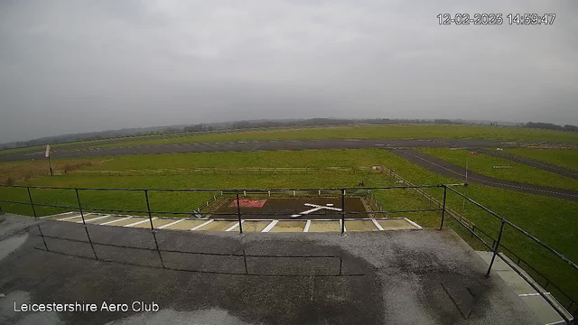 A webcam view of an outdoor airport scene on a cloudy day. In the foreground, there is a railing along the edge of a building. Below the railing, a tarmac area features a white 'X' marking on the ground, indicating a possible landing zone. The background shows a large green field bordered by a fence, and an asphalt runway extending into the distance. There is a hint of rolling hills or trees beyond the runway, and the sky appears overcast.