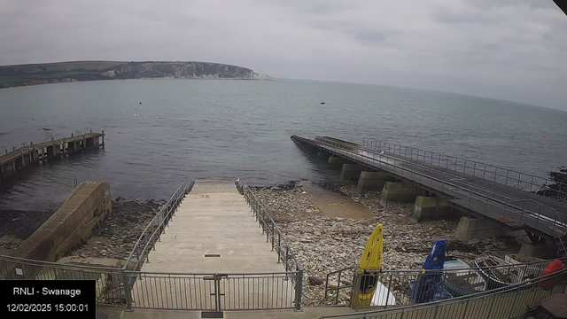 A view of the coastline featuring a calm sea under a cloudy sky. There is a stone and concrete ramp leading down to the water, bordered by a rail fence. On the right, a wooden jetty extends into the sea. Two kayaks, one yellow and one blue, are stored near the ramp. The rocky shoreline is visible, and a distant cliff can be seen in the background on the left side of the image.