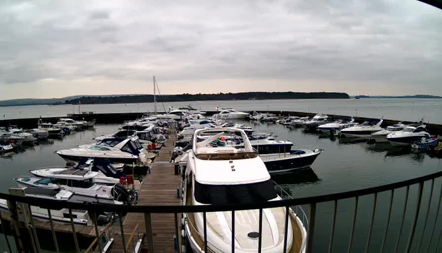 A view of a marina filled with various boats docked in calm water. The scene is set under a cloudy sky, with a large wooden pier visible in the foreground. Several boats, including powerboats and sailboats, are moored closely together. In the background, there is a distant shoreline, and the water reflects the overcast sky. Some boats have colorful covers and accessories on them.