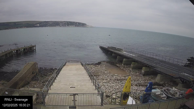 A view of a harbor with a wooden pier extending into the calm water. On the left side, there is a brick walkway leading down to the water, with a railing along its sides. To the right, a wider concrete ramp leads down to the sea. The shoreline is rocky, with several kayaks positioned near the dock, one yellow and one blue. A cliff is visible in the background, along with a cloudy sky overhead. The date and time are displayed in the lower left corner.