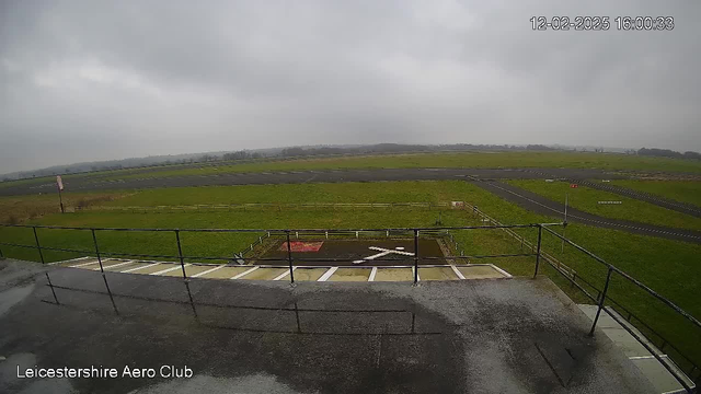 A cloudy sky is visible above a grassy area that leads to a runway at the Leicestershire Aero Club. The ground is damp, with a few distinct line markings on the tarmac. There are wooden fences lining the grassy space, and a red sign is partially visible in the distance. The scene is devoid of any aircraft or people, creating a quiet, open environment. The timestamp in the corner indicates the image was taken on December 2, 2025, at 16:00:33.