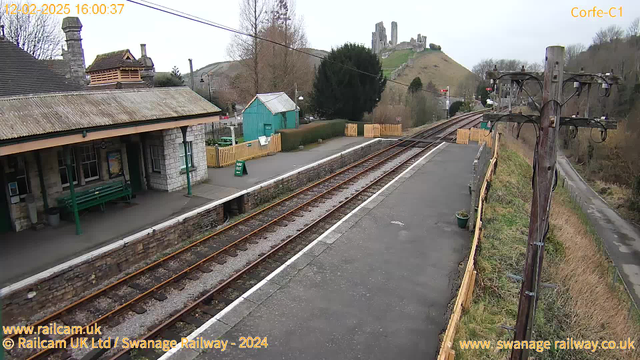 A view of a train station with a rustic stone building on the left, featuring a covered area with benches. Nearby, there is a bright green wooden shed and a sign indicating a way out. The platform has visible train tracks running towards the right, bordered by gravel. In the background, a hill with the ruins of a castle can be seen, surrounded by trees. The sky is overcast, and there is a telephone pole on the right side of the image.