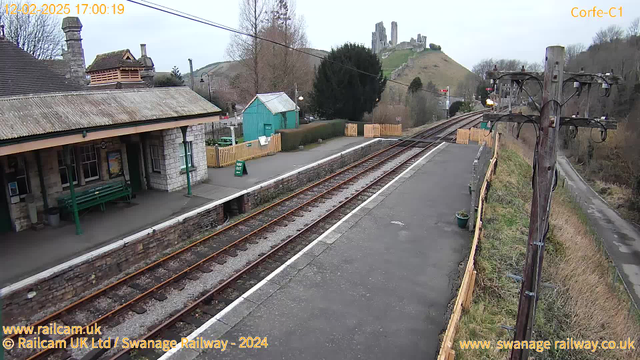 A train station platform is visible, featuring a stone building with a sloped roof and green benches. To the left, a green structure stands along with a wooden fence. Several railroad tracks run alongside the platform, which is bordered by gravel. In the background, a hillside with ruins of a castle is visible, with trees and shrubs scattered throughout. The scene is under a cloudy sky, with a power pole on the right side of the image.