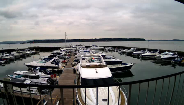 A marina filled with various boats and yachts docked in calm waters. In the foreground, a large white boat is prominently visible, with other boats of different sizes and colors surrounding it. The water is reflecting a cloudy sky, and distant green hills can be seen beyond the marina. A wooden walkway is present in the foreground, leading towards the boats, with a railing visible on the left side of the image. The atmosphere appears overcast and tranquil.