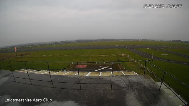 A view from a webcam situated on a building at the Leicestershire Aero Club. The scene is overcast with gray skies. In the foreground, there is a railing and a flat surface with white lines. Beyond this area is a large expanse of grass, which transitions into a dark asphalt runway. A windsock, indicating wind direction, is visible on the left side of the image. In the distance, there are blurred outlines of trees and possibly a few small vehicles or aircraft on the runway, all under a uniform gray sky.