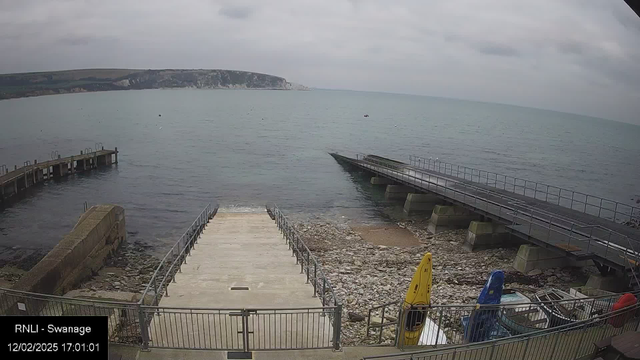 A view of a coastal area featuring a rocky shore and calm waters. In the foreground, there is a concrete ramp leading down to the water, bordered by a metal railing. To the left, a wooden pier extends into the water. On the right, several colorful kayaks are lined up, with a yellow kayak prominently visible. The background shows a slight hill with grassy slopes and a cloudy sky above. The scene conveys a tranquil, seaside atmosphere.